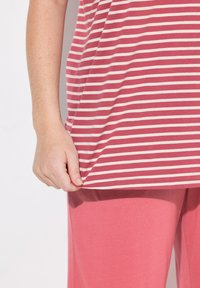 Camiseta a rayas de tela suave en rosa claro y blanco, con mangas cortas y un dobladillo redondeado, combinada con pantalones rosa a juego. Una mano sostiene el borde de la camiseta.