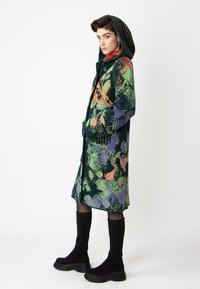 Multicolored wool coat with a hood, featuring a green base and abstract patterns in orange, purple, and black. Buttons and textured sleeves.
