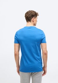 Άνδρας με κοντά καστανά μαλλιά, φορώντας μπλε κοντομάνικη μπλούζα και ανοιχτό γκρι παντελόνι, στέκεται με την πλάτη προς το μέρος μας πάνω σε  απλό φόντο.