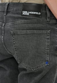 Sive traperice imaju crnu kožnu oznaku s utisnutim natpisom "KARL LAGERFELD JEANS". Uključuje plavi detalj na stražnjem džepu i standardno šivanje.
