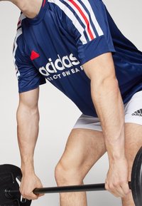 Manlig idrottare som bär en blå Adidas sporttröja och vita shorts, lyfter en viktstång med båda händerna i en gymmiljö.