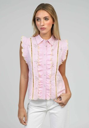 Femme portant un chemisier rose sans manches avec des volants et des détails en dentelle, associé à un pantalon blanc, debout devant un fond gris uni.