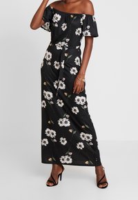 Maxi šaty bez ramínek černé barvy s květinovým vzorem v bílé a žluté barvě, páskem v oblasti pasu a splývavou tkaninou, doplněné černými lace-up podpatky.