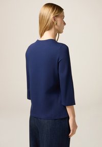 Donna con lunghi capelli biondi che indossa orecchini d'oro a forma di ventaglio, un top blu navy a maniche tre quarti e pantaloni in denim scuri, di spalle.