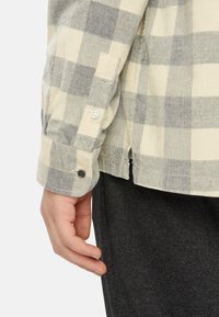 Chemise à manches longues à carreaux beige et gris avec des poignets boutonnés, présentant un tissu texturé et des fentes latérales au niveau de l'ourlet.