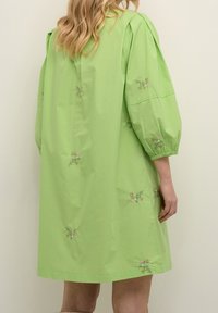 Femme portant une robe vert citron mi-longue avec des manches bouffantes et une petite broderie florale, debout devant un fond clair uni.