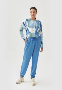 Blauer Sweatshirt mit abstraktem Muster und langen Ärmeln, kombiniert mit blauen Jogginghosen und cremefarbenen Sneakers. Glattes Baumwollmaterial.