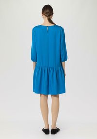Blaues knielanges Kleid mit ¾-Ärmeln und geraffter Taille. Mit einem Schlüsselaugen-Detail im Rücken und einer glatten Stofftextur.
