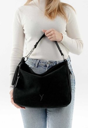 Woman wearing white sweater and blue jeans holds a large black handbag with a detachable handle and zipper details.