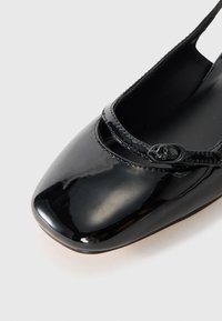 Ballerine in vernice nera con punta quadrata, dotate di una sottile fascetta e un piccolo dettaglio a fibbia. Accenti testurizzati attorno all'apertura.