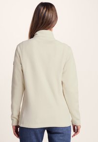 Fleece pullover in cream with a high collar, long sleeves, and a side logo accent. Soft texture and relaxed fit, suitable for layering.