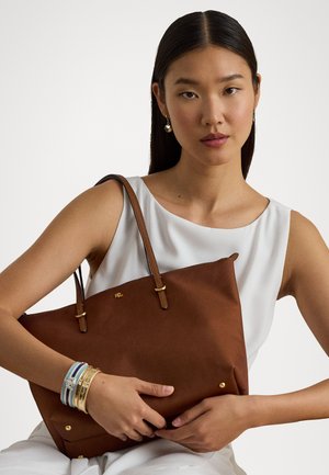 Καφέ τσάντα ώμου με δερμάτινες λαβές, χρυσές λεπτομέρειες και απαλό υφασμά. Μοντέλο φορώντας ένα λευκό μπλουζάκι, κρατώντας την τσάντα.