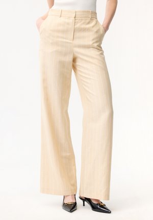 Beige High-Waist-Hose mit weitem Bein und dezenten Nadelstreifen, kombiniert mit schwarzen spitzen Pumps und weißem ärmellosem Oberteil. Hände in den Taschen.
