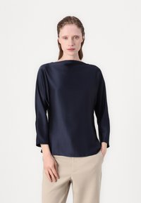 Marine satinbluse med lange ermer, båthals og jevn tekstur. Brukt sammen med lyse beige bukser. Enkel, moderne design.