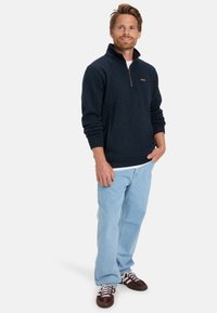 Navy fleece pullover with a half-zip collar, kangaroo pocket, and a brown leather logo patch, paired with light blue denim jeans.