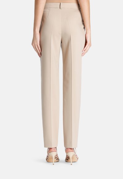 Pantalons beige ajustés avec une jambe droite, un devant plissé et une texture lisse. Associés à des sandales à talons hauts transparentes.