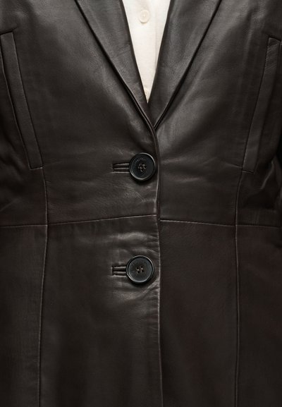 Blazer en cuir marron foncé boutonné sur un haut en maille blanche avec deux boutons noirs visibles au centre devant.