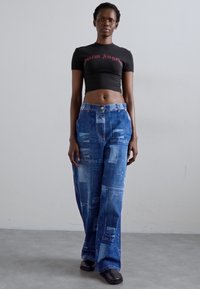 Black cropped shirt with "Palm Angels" text, paired with loose blue jeans featuring graphic prints. Model wears black shoes.