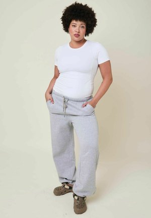 Personne aux cheveux bouclés, debout, portant un t-shirt blanc ajusté, un pantalon de survêtement gris ample et des sabots marron à enfiler avec des sangles. Mains dans les poches.