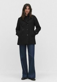 Musta kaksirivinen takki suurilla napilla, jossa on sivutaskut ja sileä, jämäkkä kangas. Yhdistetty leveälahkeisiin farkkuihin.