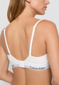 Hvit bh med justerbare stropper, en glatt overflate og et merket elastisk bånd med "Calvin Klein" i svart.