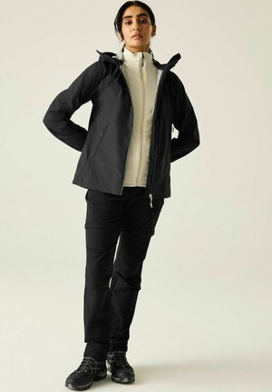 Woman wearing black outdoor jacket, black pants, and black hiking shoes, standing with hands behind back on light background.