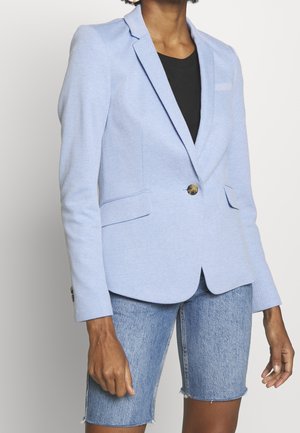 Vrouw draagt een lichtblauw colbert over een zwart topje, gecombineerd met knielange blauwe denim shorts, staand tegen een lichte achtergrond.