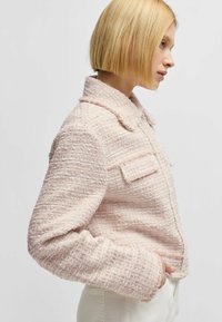 Vaaleanpunainen tweed-takki, jossa on teksturoitu kangas, kaulus ja kaksi etutaskua. Sisältää lyhennetyn muotoilun ja pehmeät, rispaantuneet reunat.