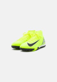 Zapatos de fútbol de color amarillo neón con acentos en negro, que cuentan con un diseño de tobillo alto y una parte superior texturizada, diseñados para superficies de césped artificial.
