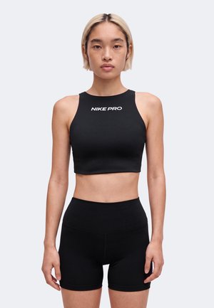 Jeune personne aux cheveux courts blonds portant un crop top noir Nike Pro et un short noir taille haute sur un fond uni.