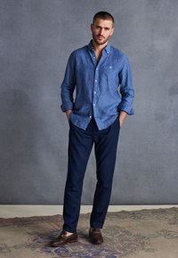 Cămașă din in bleu deschis, pantaloni navy și mocasini maro închis. Cămașa are mâneci suflate și un singur buzunar pe piept.