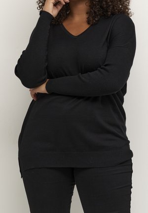 Femme aux cheveux bouclés portant un pull noir à manches longues avec col en V et un pantalon noir, les bras croisés et la main près du visage.