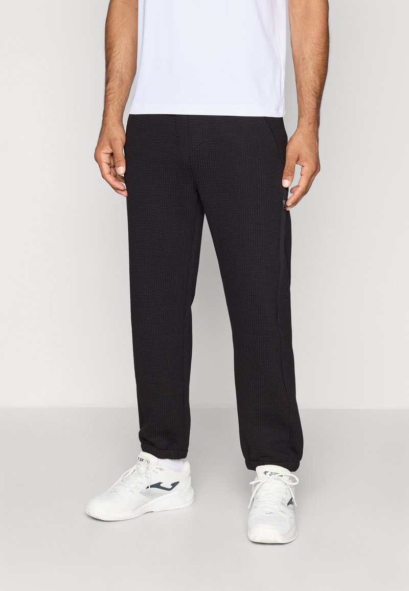 Black ribbed sweatpants made of textured fabric, featuring side pockets and a fitted design, paired with white athletic shoes.