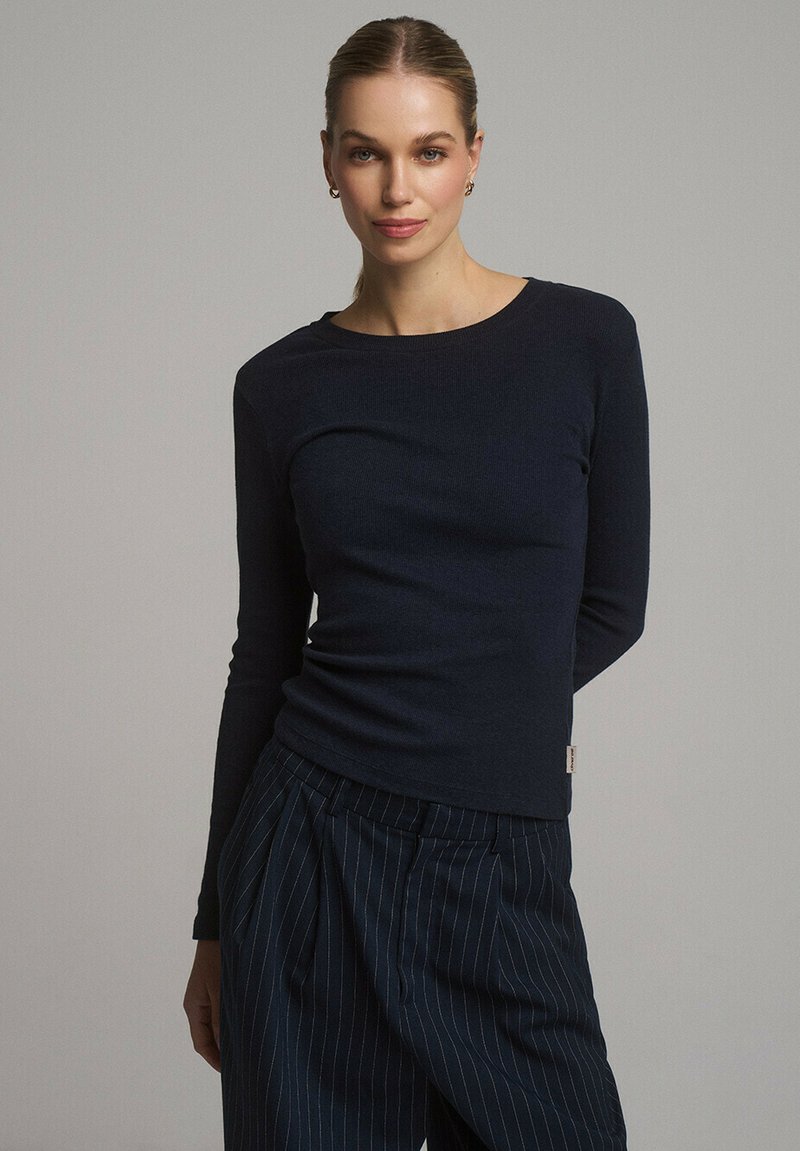 Granatowa bluzka z długim rękawem w prążkowanej strukturze z okrągłym dekoltem. Połączona ze spodniami w kant z plisami, podkreślającymi dopasowaną sylwetkę.