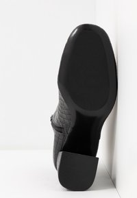 Bottine noire avec un motif crocodile texturé, un petit talon et une fermeture éclair sur le côté. Semelle lisse et design à bout arrondi.