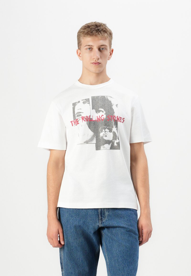 Ung mand iført en hvid T-shirt med sort-hvide billeder af Rolling Stones-bandet og rød tekst, parret med blå jeans, stående mod en hvid baggrund.