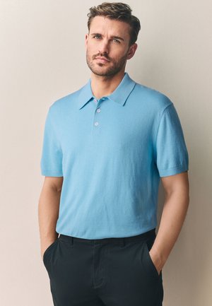 Lys blå kortærmet polo shirt med opfoldet krave og tre knapper, parret med mørke bukser, stående mod en neutral baggrund.