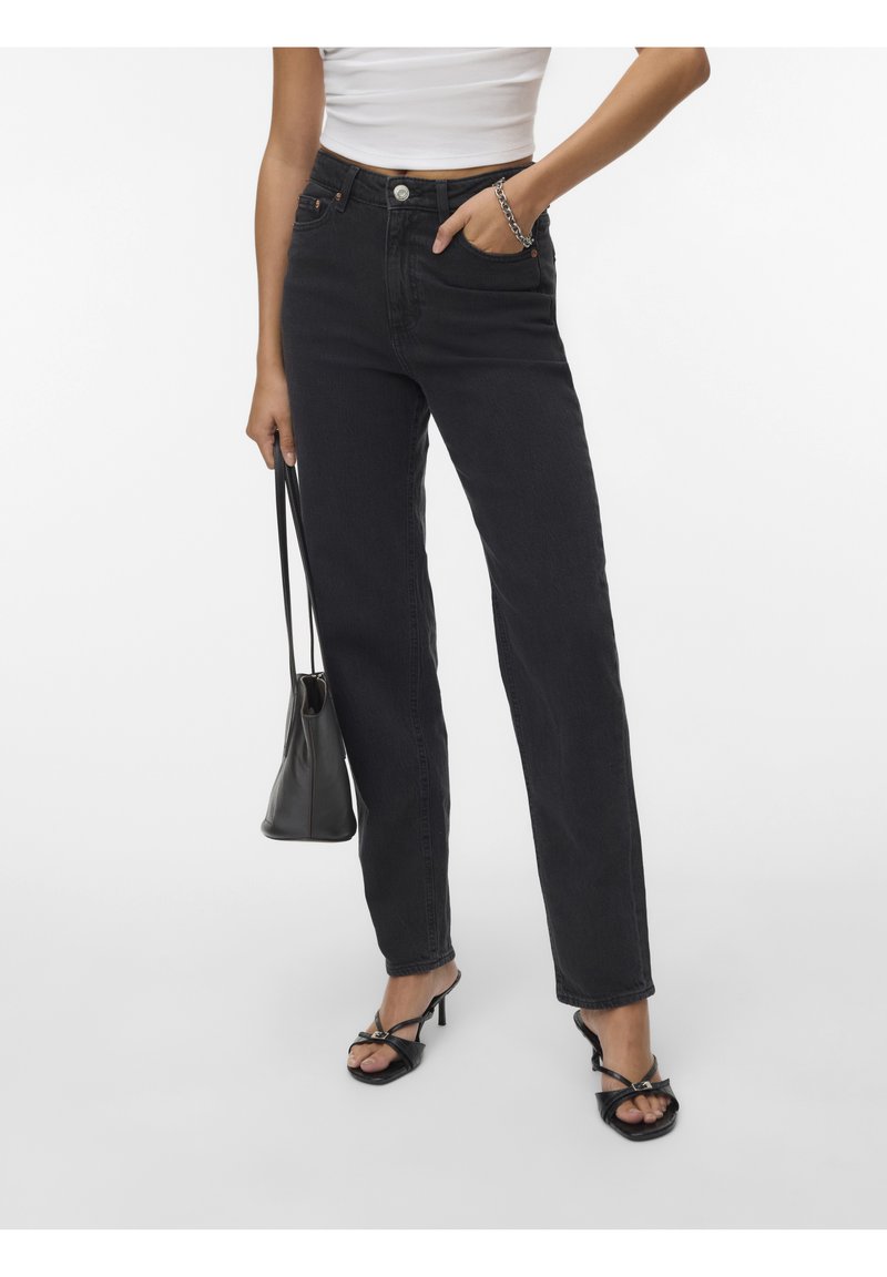 Jeans noirs taille haute à coupe droite, en denim, avec poches avant et fermeture par bouton. Portés avec des talons noirs à lanières.