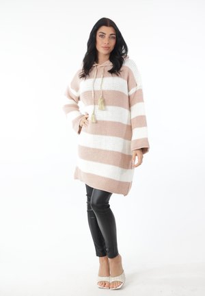 Frau steht mit der Hand in der Hüfte, trägt ein beige-weiß gestreiftes Strick-Kapuzenkleid, schwarze Leggings und weiße offene Absätze vor weißem Hintergrund.