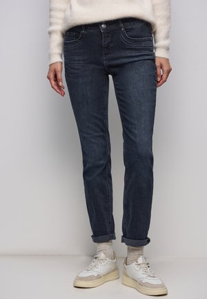 Dunkelblaue Slim-Fit-Denimjeans mit Vordertaschen, aufgekrempelten Bündchen und dezenten Verwaschungen. Kombiniert mit weißen und cremefarbenen Sneakers.