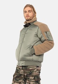 Grøn polstret jakke med beige kunstpelsdetaljer, høj krave og frontlynlås. Har sidelommer og en lomme på armen til praktisk brug.