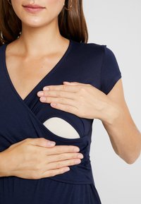 Kvinna visar hur man öppnar en marinblå amningsklänning med en krämfärgad innerfoder synlig under.