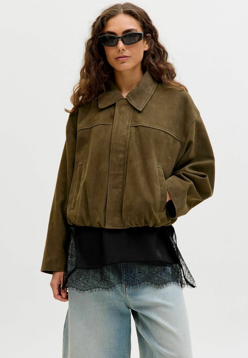 Femme aux cheveux longs et bouclés portant une veste oversize vert olive, un chemisier noir orné de dentelle, un jean bleu clair et des lunettes de soleil étroites noires.