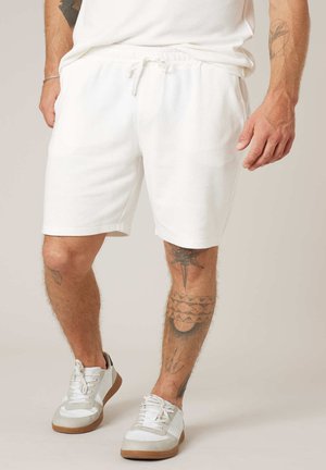 Homme portant un short blanc à cordon, des baskets blanches et une chemise blanche, avec des tatouages visibles sur les bras et les jambes, sur un fond uni.