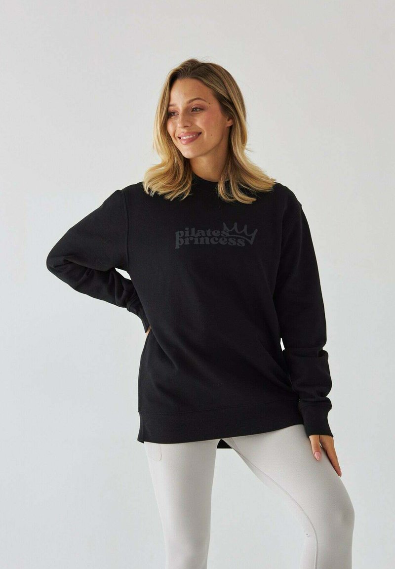 Czarny sweatshirt z napisem "księżniczka pilatesu" oraz logo korony. Długie rękawy, luźny krój, zestawiony z jasnymi legginsami.