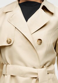 Μπεζ καμπαρντίνα με φαρδιούς γιακάδες, διπλή σειρά κουμπιών, ζώνη στη μέση, φορεμένη πάνω από μαύρο τοπ.