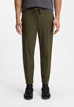 Mann trägt olivgrüne, schmal zulaufende Hosen mit Kordelzug an der Taille, schwarze Sneakers und schwarze Socken, steht vor einem schlichten weißen Hintergrund.