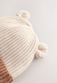 Fehér kötött sapka bordás mintával, tetején két kerek pomponnal és az alján kontrasztos barna szegéllyel.