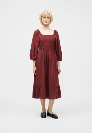Femme aux cheveux blonds bouclés portant une robe midi bordeaux à manches longues et des ballerines noires, debout devant un fond blanc uni.