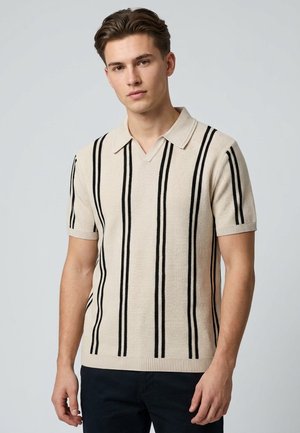 Junger Mann trägt ein beiges kurzärmliges Strickshirt mit schwarzen vertikalen Streifen und Kragen und steht vor einem schlichten Hintergrund.